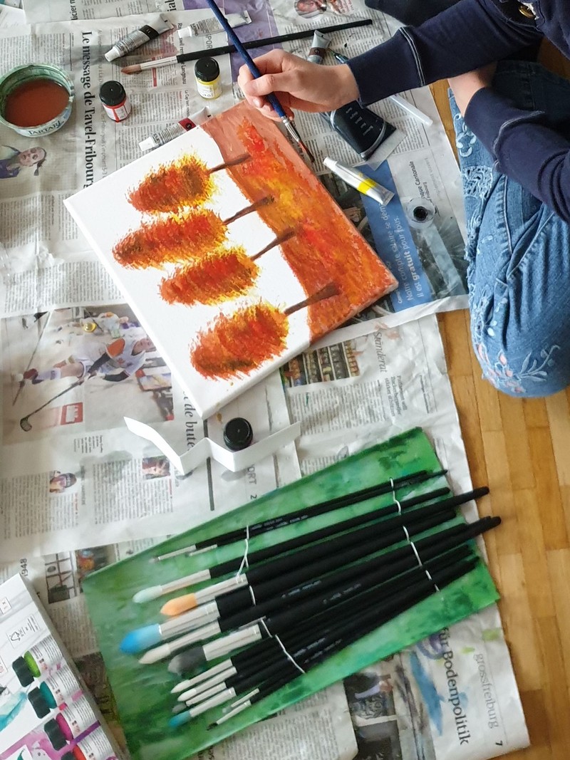 Réka painting autumn trees on a small canvas, brushes and paints spread out on the floor